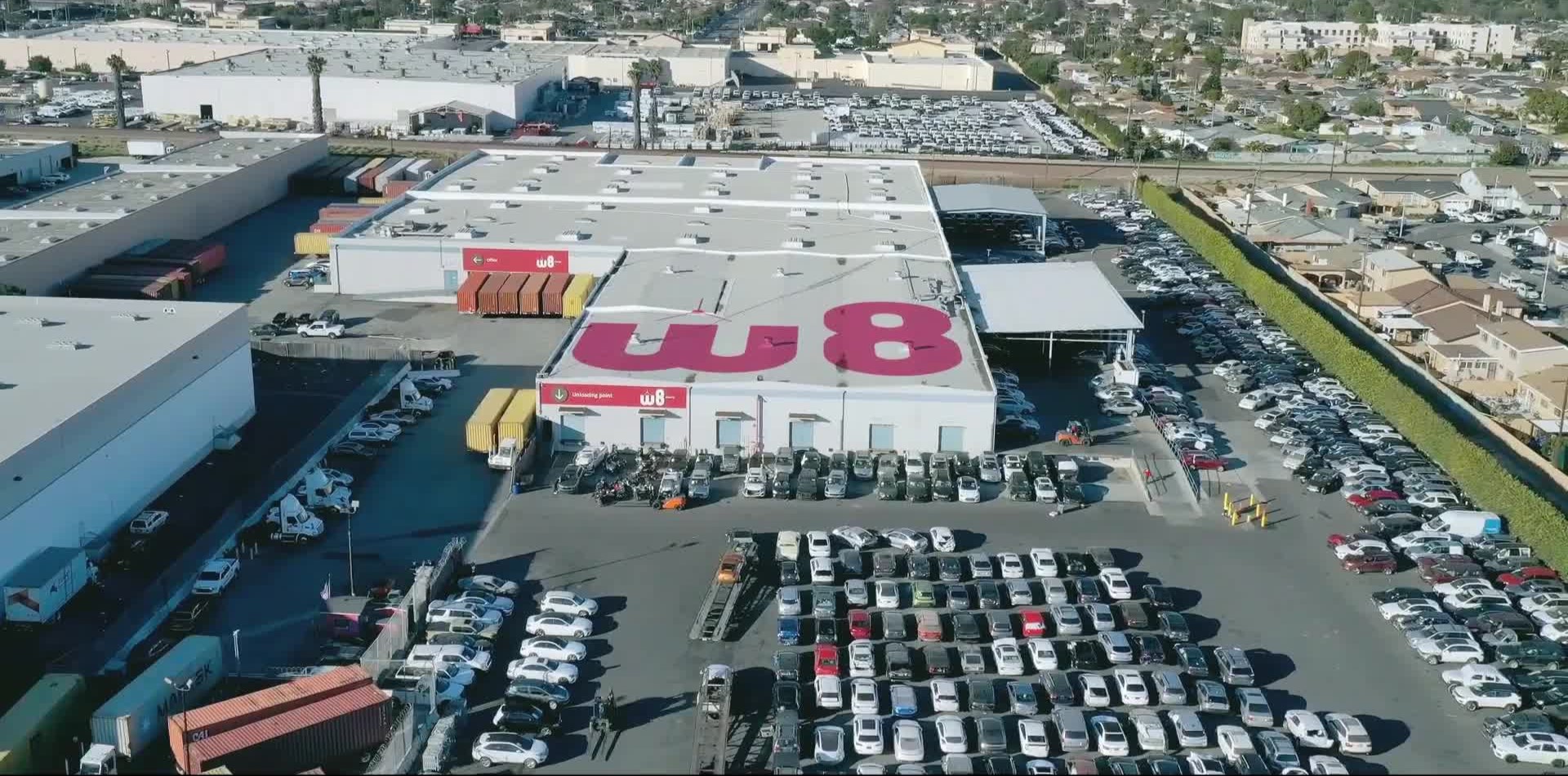 У центрі фотографії склад із великим логотипом W8 на даху та меншими логотипами над кількома входами. Навколо складу розташоване багато автомобілів, припаркованих на парковці.